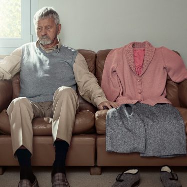 Kampagnenfoto: Mann sitzt vereinsamt auf einem Sofa. Neben sich hat er Frauenkleider präpariert. Diese symbolisieren die Abwesenheit einer Partnerin.
