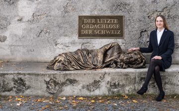 Eine Heilsarmee-Offizierin sitzt neben einer Statue eines Obdachlosen. Oberhalb der Statue steht an der Wand der Schriftzug 